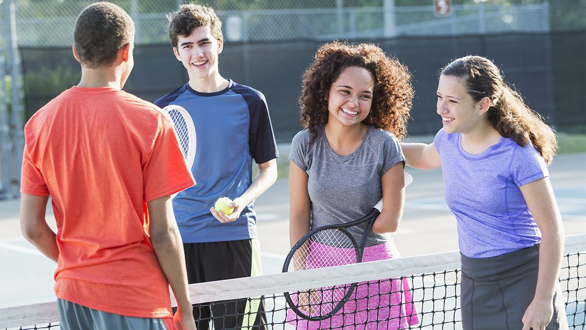 A group of four multiracial teenage girls and boys playing mixed doubles tennis.  They are standing at the net, relaxed, talking and smiling, holding tennis rackets.  The girl in the gray shirt and pink skirt is a physically challenged amputee, 17 years old, mixed race African American.